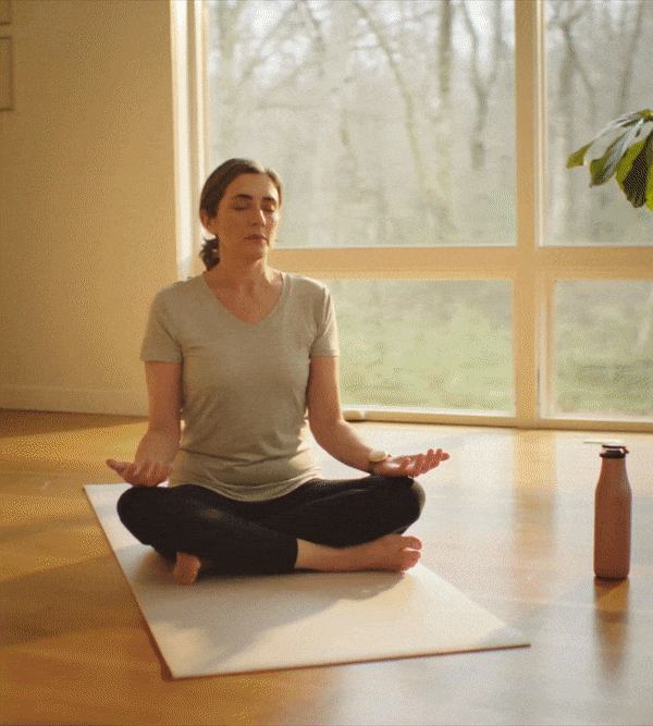 Woman meditating on yoga mat by sunny window wearing the Embr Wave bracelet.